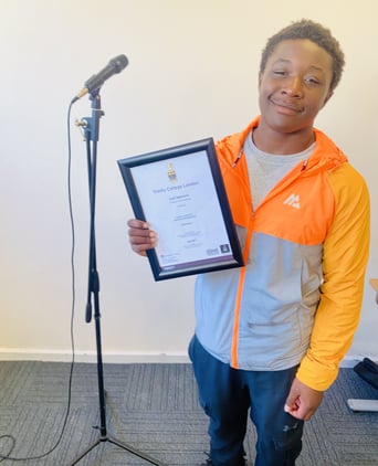Black teenager wearing blue joggers, a grey t-shirt and orange and grey hooded jacket smiles as he stands next to a microphone holding his framed ACMD certificate