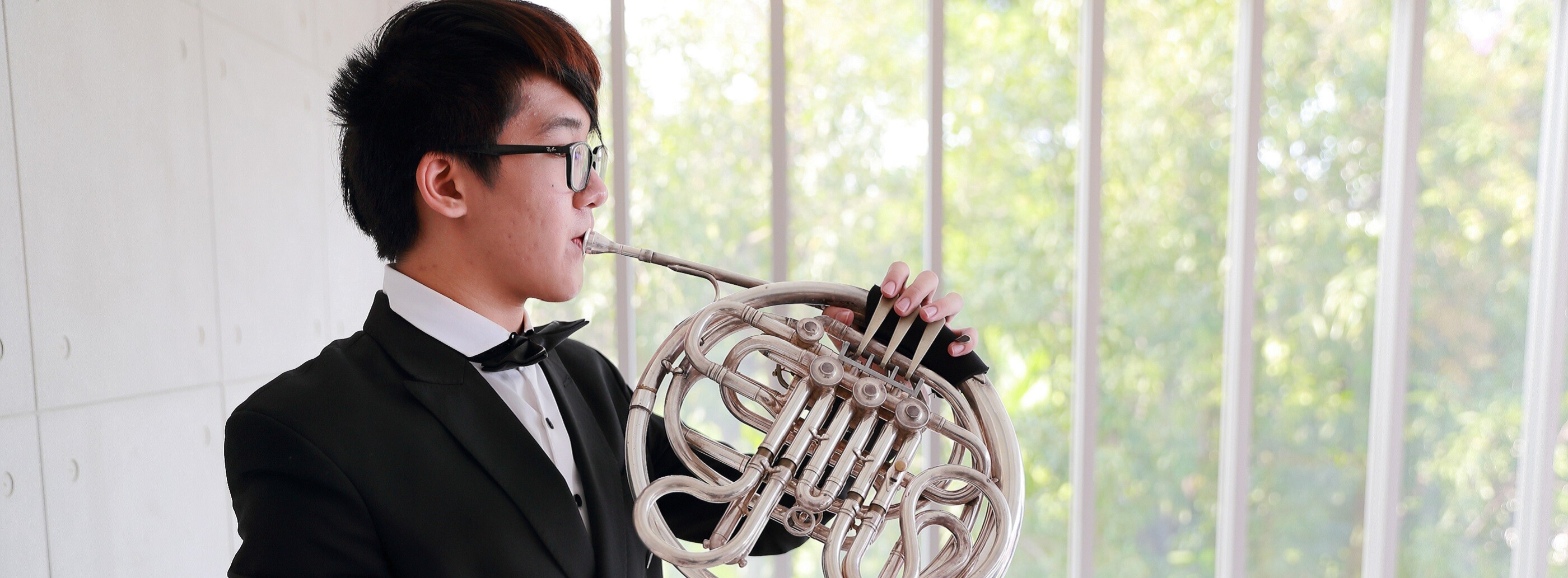 Young man with black hair and black glasses, wearing a tuxedo, looks out of the window while playing a silver French horn