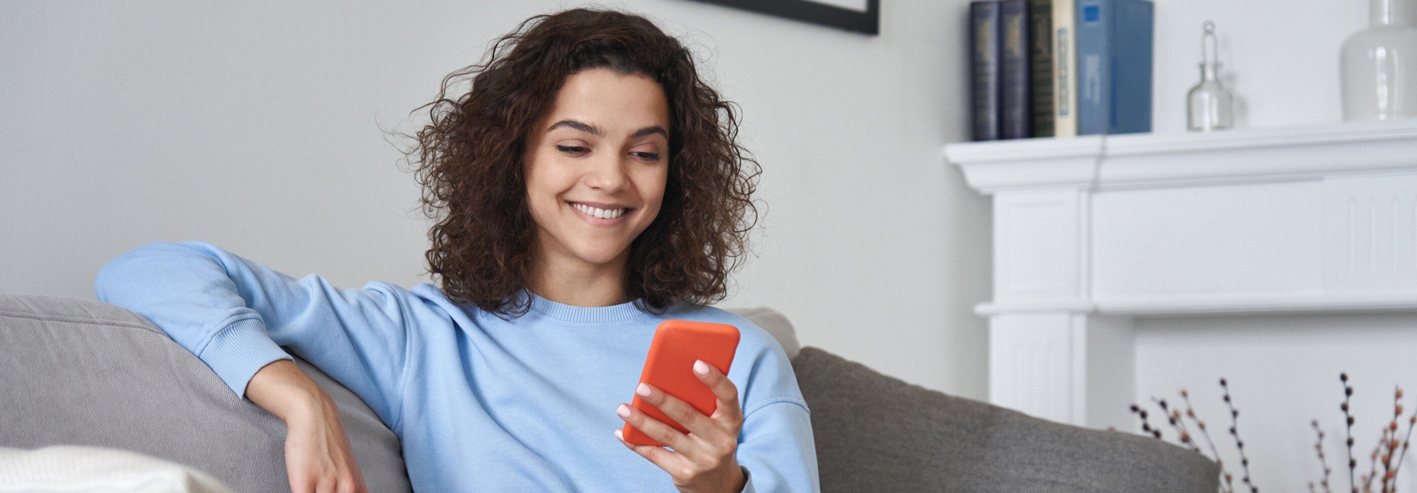 Woman with brown curly hair sits on grey sofa looking at her orange smartphone