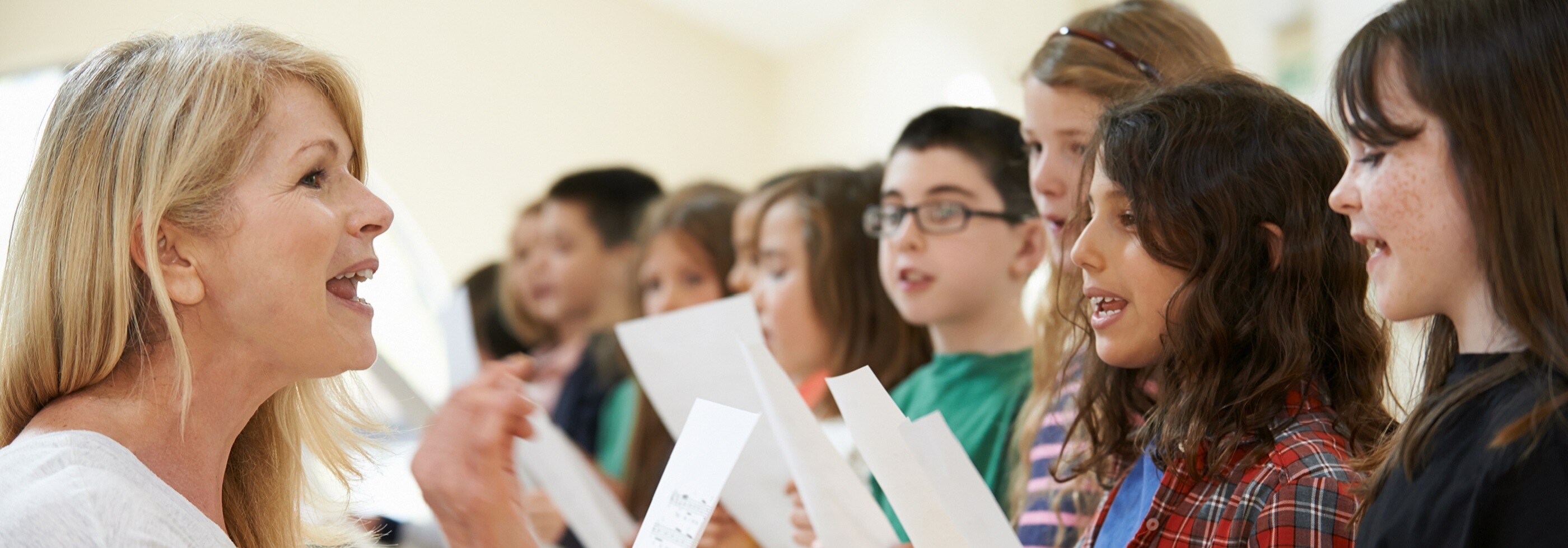 Woman with long blonde hair and white top teaching group of children to sing