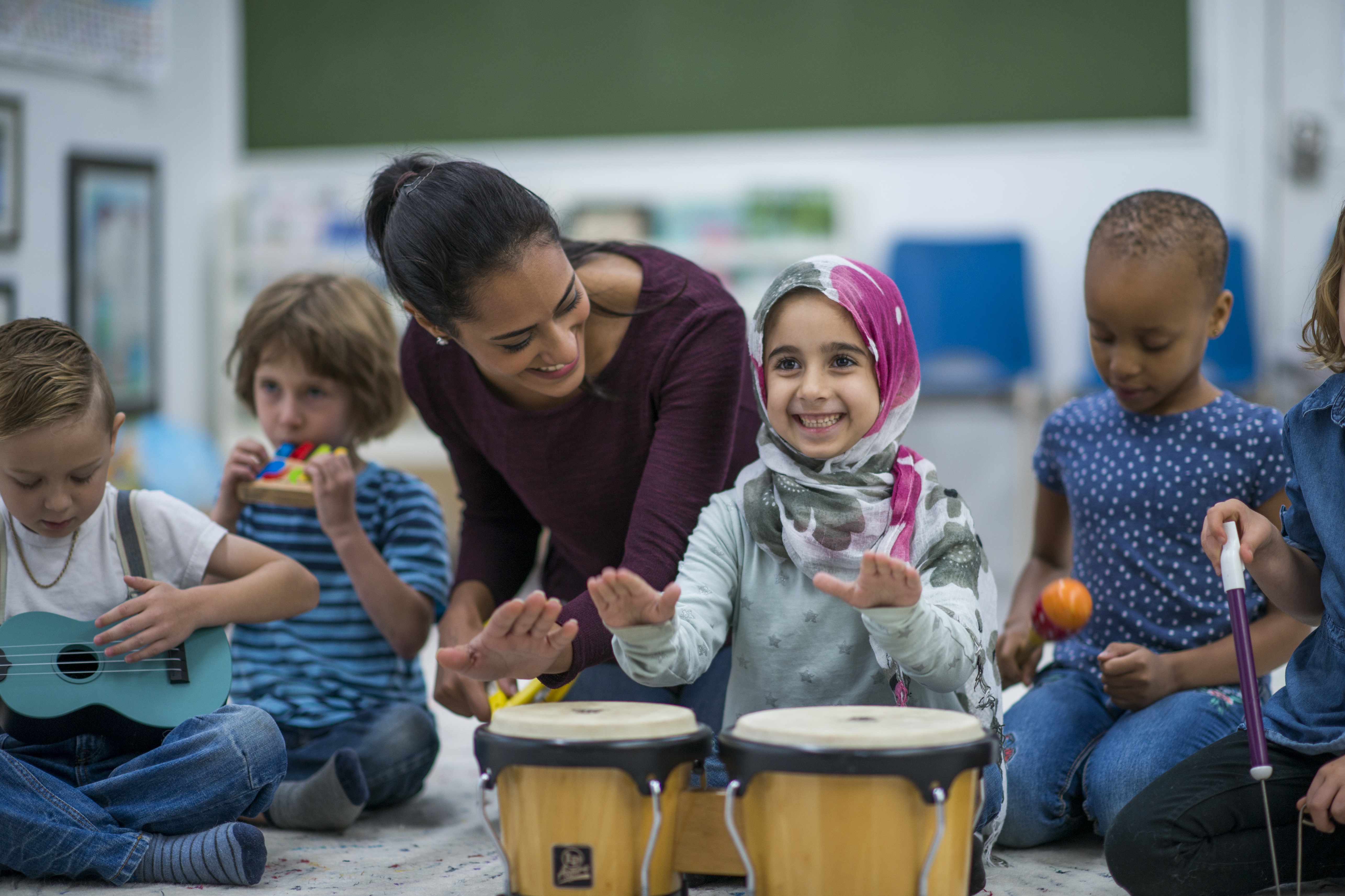 A young girl in a headscarf in a percussion class. She is playing the bongos with her female teacher alongside her.