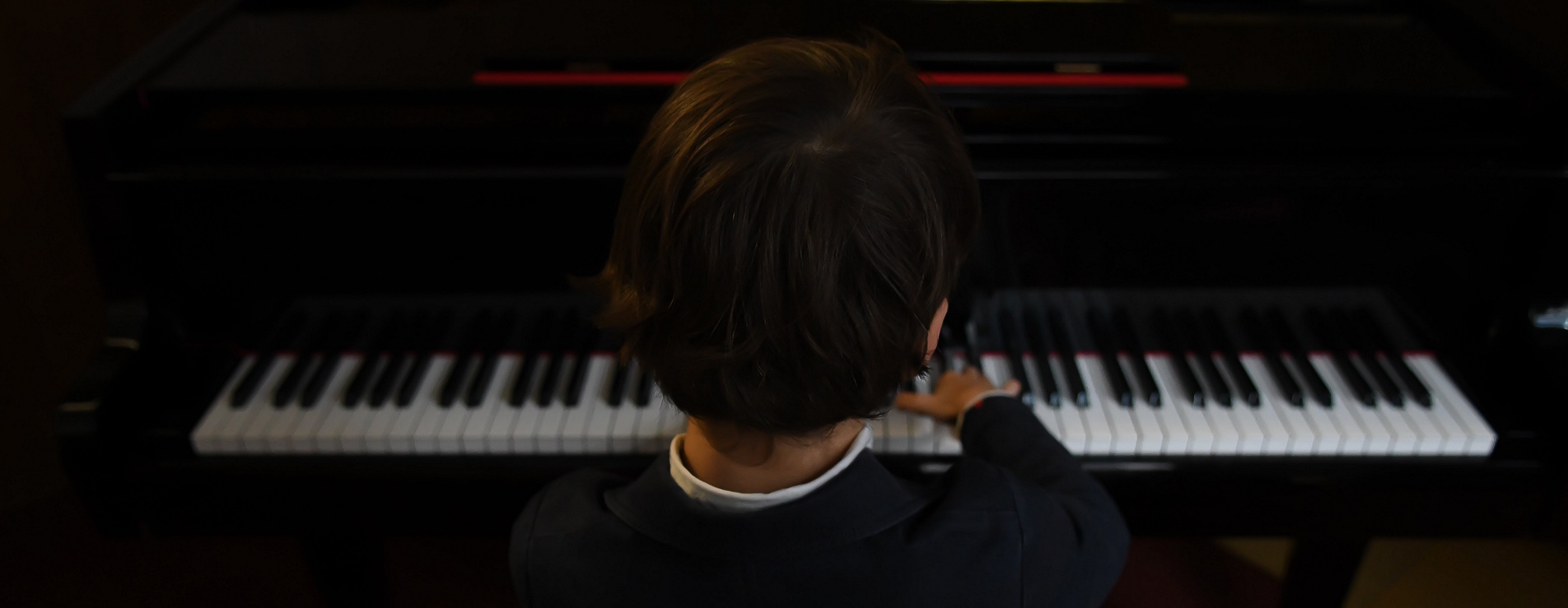Behind the head shot of a little boy with brown hair, white collar and blue jacket playing the piano
