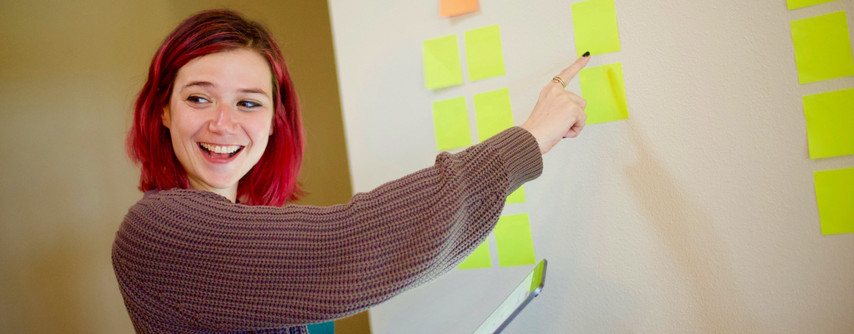 Lady with bright red hair smiling and pointing to a white board with post-it notes on it