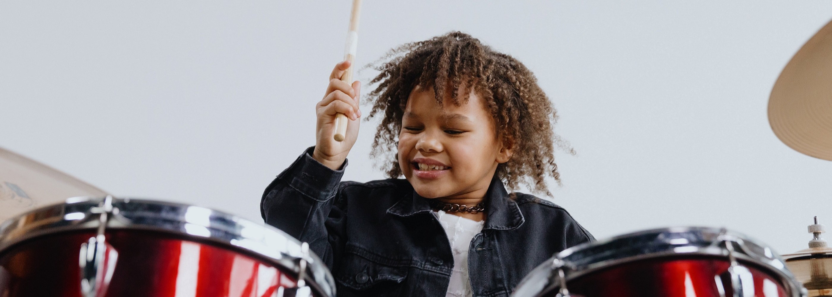 Young girl with brown, afro hair, white t-shirt, black denim jacket and chain necklace holding up a drumstick as she plays a red drum kit