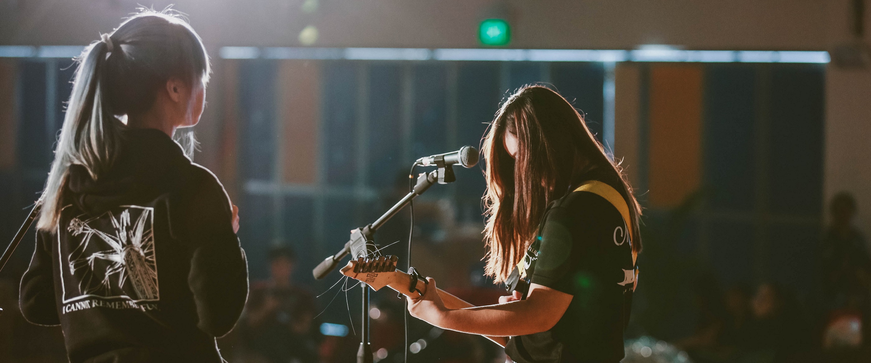 A shot taken from the back of a stage where two teenage girls are performing. One is in front of the microphone, playing guitar.