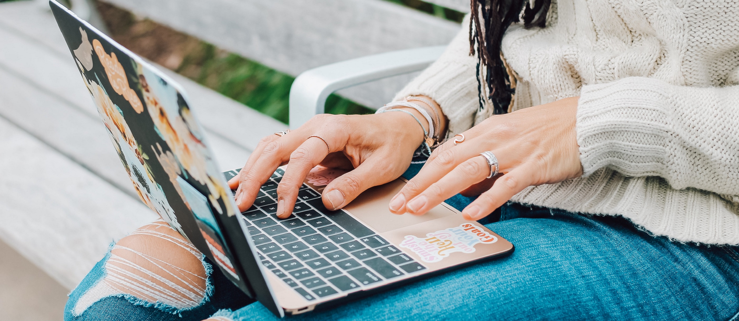 Laptop resting on the lap of a white person wearing ripped jeans and a cream jumper. The user's hands are resting on the laptop and are adorned with rings and bracelets.