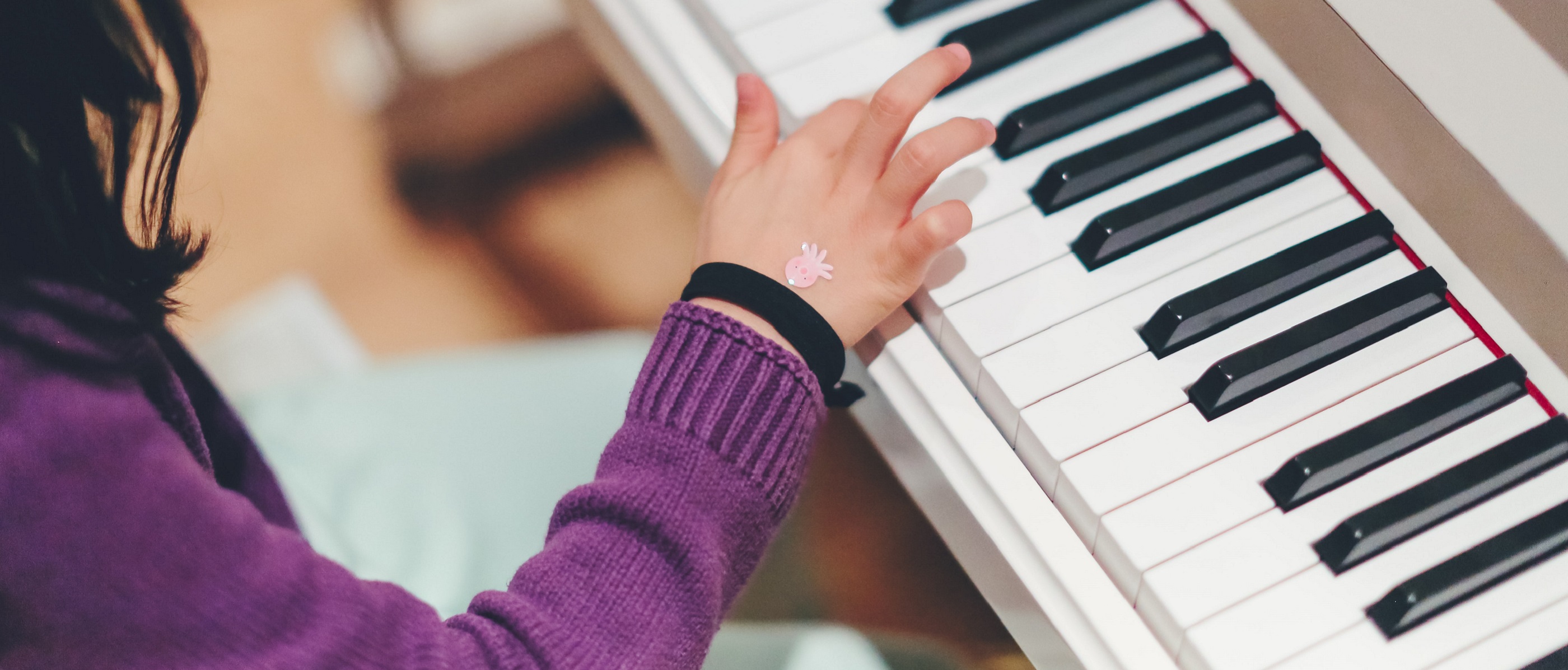 A child's right arm and hand as they press a key on a white piano. They wear a purple jumper, a black wristband and a pink octopus sticker on their hand.